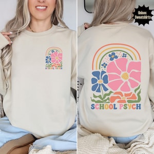 May include: A beige sweatshirt featuring a rainbow and flower design. The text "SCHOOL PSYCH" is printed on the front and back of the sweatshirt.