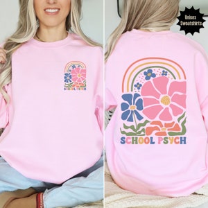 May include: Pink unisex sweatshirt featuring a rainbow and flower design. The text "SCHOOL PSYCH" is printed on the sweatshirt.