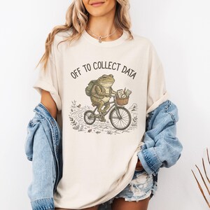 May include: Cream-colored t-shirt featuring a whimsical illustration of a frog riding a bicycle with a basket. The frog wears a backpack and is riding on a path. The text "OFF TO COLLECT DATA" is printed above the frog. The shirt has short sleeves.