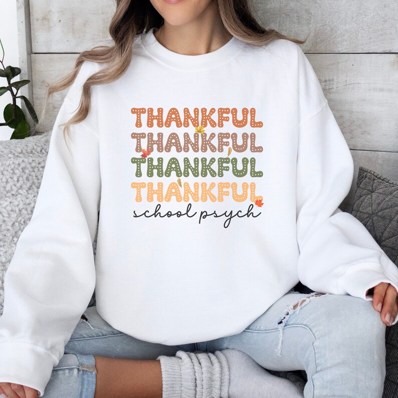 Puede incluir: Sudadera blanca con el texto "THANKFUL" repetido cuatro veces en diferentes colores y "school psych" escrito debajo. El texto est&aacute; decorado con peque&ntilde;os dise&ntilde;os de hojas.