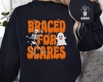 May include: Black sweatshirt with an orange graphic that says "Braced for Scares" with a skeleton and ghost. The back of the sweatshirt has a white spider web and tooth graphic with the name "Sarah".