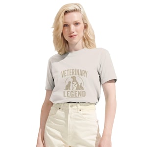 Può includere: T-shirt grigio chiaro con la scritta "VETERINARY LEGEND" in beige. La grafica include un cane, un gatto e un coniglio, insieme a un simbolo medico. La maglietta è indossata da una persona con i capelli biondi ed è abbinata a jeans chiari.