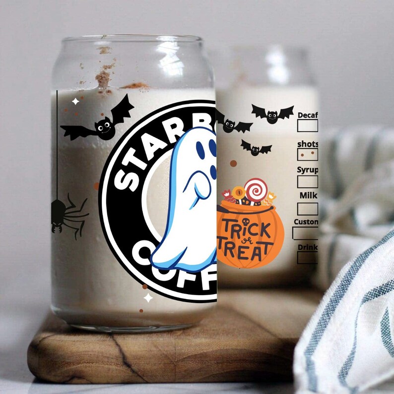 May include: A clear glass can with a black and white Starbucks logo and a ghost graphic. The can has a white background with black bats and a spider. The text "Trick or Treat" is on the can with a pumpkin graphic. The can is on a wooden surface.