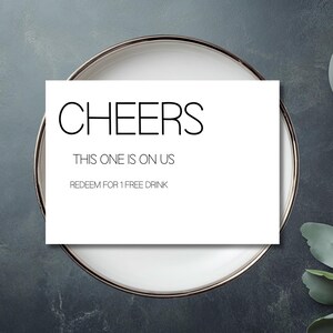 Könnte beinhalten: Eine weiße Karte mit dem Wort "CHEERS" in fetter, schwarzer Schrift. Darunter steht der Text "THIS ONE IS ON US" und "REDEEM FOR 1 FREE DRINK". Die Karte liegt auf einem weißen Teller mit silbernem Rand.