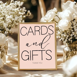 Puede incluir: Un letrero enmarcado en dorado con las palabras "CARDS and GIFTS" en escritura negra sobre un fondo rosa claro. El letrero incluye las palabras "THANK YOU" en la parte inferior. Flores blancas en jarrones de cristal en el fondo.