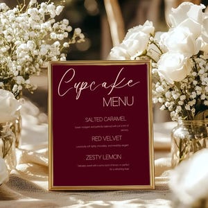 May include: A framed cupcake menu with a burgundy background and gold trim. The menu features the word "Cupcake" in elegant script, followed by "MENU." Flavours listed include Salted Caramel, Red Velvet, and Zesty Lemon. White flowers are in the background.