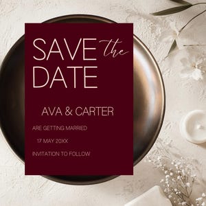 Puede incluir: Una tarjeta burdeos de Save the Date con texto blanco, con los nombres "AVA & CARTER" y la fecha de la boda "17 MAYO 20XX". La tarjeta está colocada sobre un plato de color bronce, con flores blancas y una vela en el fondo.