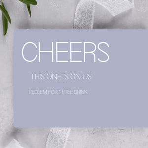 Puede incluir: Una tarjeta de regalo rectangular azul claro con la palabra "CHEERS" en blanco, con una fuente estilizada. Debajo, la tarjeta dice "THIS ONE IS ON US" y "REDEEM FOR 1 FREE DRINK". La tarjeta está sobre un fondo gris claro texturizado con una cinta blanca y vegetación.