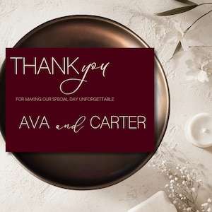 Könnte beinhalten: Eine weinrote Dankeskarte mit weißem Text, der "THANK you" und "FOR MAKING OUR SPECIAL DAY UNFORGETTABLE" sowie die Namen "AVA and CARTER" lautet. Die Karte liegt auf einem dunklen Bronzeteller.