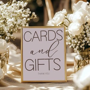 May include: A framed sign with the words "CARDS and GIFTS" in dark gray script. The sign has a gold frame and a light purple background. The words "THANK YOU" are at the bottom. The sign is on a table with flowers.
