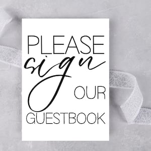 May include: A white rectangular sign with the words "PLEASE sign OUR GUESTBOOK" in black. The word "sign" is in a cursive font. A white lace ribbon is draped across the top and bottom of the sign.