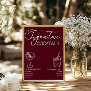 May include: A framed burgundy sign with gold trim displays "Signature Cocktails" in elegant script. Below, two cocktail illustrations are featured with their ingredients: Blushing Bride Spritz and Old Fashioned Twist. A vase of white flowers sits nearby.
