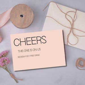 May include: A blush pink gift card with the word "CHEERS" in black lettering. The card also reads "THIS ONE IS ON US" and "REDEEM FOR 1 FREE DRINK". The card is next to a wooden spool, a pink ribbon, a bundle of dried flowers, and a cream-colored envelope tied with twine.