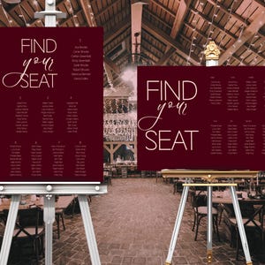 May include: Two burgundy seating chart signs with white text that read "FIND your SEAT". The signs are on easels in a rustic venue. Each sign lists names and table numbers for guests. The background is a blurred view of a reception.