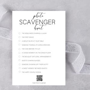 May include: A white card with the words "photo scavenger hunt" in elegant script. The card lists prompts for wedding photos, including "the first dance" and "the happy couple kissing." A QR code is at the bottom. A white ribbon is in the background.