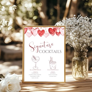 May include: A framed sign with the words "Signature Cocktails" in elegant script. The sign features watercolor hearts and illustrations of two cocktails: "Blushing Bride" and "Old Fashioned". The sign is in a gold frame.
