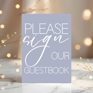 Puede incluir: Un letrero rectangular con el texto "PLEASE sign OUR GUESTBOOK" en letras blancas sobre un fondo lila claro. El letrero es probablemente para una boda o evento.