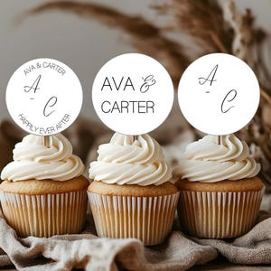 Może przedstawiać: Trzy babeczki z białym lukrem i okrągłymi topperami. Toppery mają imiona "Ava & Carter" i inicjały "A - C" w czarnym skrypcie. Jeden topper mówi również "Happily Ever After". Babeczki leżą na tkaninie w neutralnym kolorze.