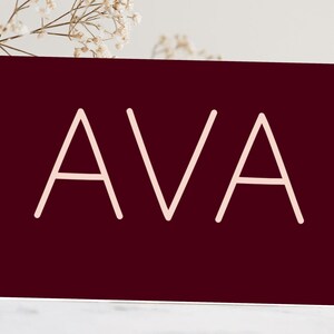 May include: A burgundy card featuring the name "AVA" in a pale pink, minimalist font. The card is set against a neutral background with delicate floral accents, suggesting a personalized gift or stationery item.