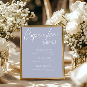 Puede incluir: Un menú de cupcakes enmarcado en oro con un fondo azul claro. El menú enumera tres sabores de cupcakes: Caramelo salado, Terciopelo rojo y Limón con sabor. Flores blancas y vegetación rodean el menú, creando una estética suave y elegante.