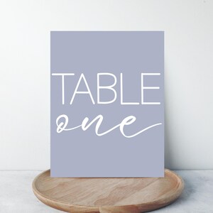 Puede incluir: Un letrero rectangular lila claro con las palabras "TABLE ONE" en blanco. El letrero está sobre una bandeja de madera. El texto está en una fuente simple y elegante. Ideal para bodas.