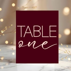 Puede incluir: Una tarjeta de número de mesa burdeos con las palabras "TABLE ONE" en una fuente blanca y elegante. La tarjeta se encuentra sobre un fondo borroso con iluminación suave y cálida y elementos decorativos.
