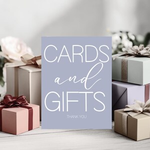 Puede incluir: Un letrero lila claro con las palabras "CARDS and GIFTS" en escritura blanca. Varias cajas de regalo envueltas en tonos rosa, verde y beige están dispuestas alrededor del letrero, cada una adornada con cintas decorativas.