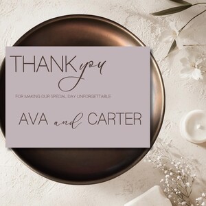Puede incluir: Una tarjeta de agradecimiento lila con las palabras "THANK you" en una fuente elegante. La tarjeta también incluye el texto "FOR MAKING OUR SPECIAL DAY UNFORGETTABLE" y los nombres "AVA and CARTER". La tarjeta está sobre un plato de color bronce.