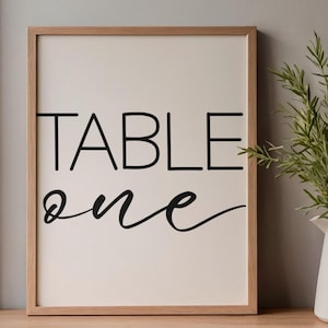 May include: A framed sign with the words "TABLE ONE" in black lettering. The word "TABLE" is in a bold, sans-serif font, while "one" is in a cursive script. The frame is made of light-colored wood.