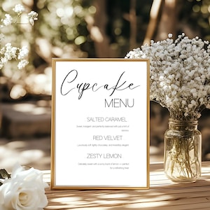 May include: A gold-framed "Cupcake Menu" sign with black script. The menu lists Salted Caramel, Red Velvet, and Zesty Lemon cupcakes. The sign is on a wooden surface, with white flowers and greenery in the background.