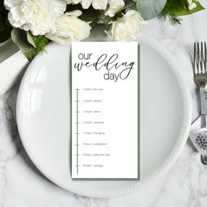 May include: A white wedding day timeline card with black text, including the words "Our Wedding Day." The schedule lists times for the vows, photos, dinner, speeches, first dance, evening food, cake cutting, and carriages.