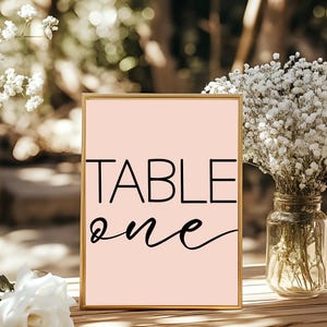 May include: A framed sign with the words "TABLE ONE" in black lettering on a light pink background. The sign has a gold frame and is displayed outdoors near flowers and a glass jar.
