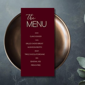Puede incluir: Una tarjeta de menú burdeos con las palabras "The MENU" en elegante escritura blanca. El menú enumera entrantes, platos principales, postres y guarniciones, incluyendo Clam Chowder y patatas fritas. La tarjeta descansa sobre un plato color bronce.