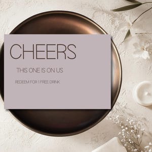 Puede incluir: Una tarjeta rectangular con la palabra "CHEERS" en letras grandes de color marrón oscuro. Debajo, el texto dice "THIS ONE IS ON US" y "REDEEM FOR 1 FREE DRINK". La tarjeta está colocada sobre un plato de color bronce.