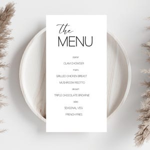 May include: A simple menu card with the words "The MENU" in black script. The menu lists starter, mains, dessert, and sides, including Clam Chowder, Grilled Chicken Breast, and Triple Chocolate Brownie. The card is set on a light wooden plate.