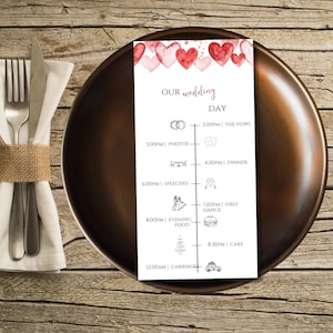 Könnte beinhalten: Eine Hochzeits-Tagesablaufkarte mit Aquarellherzen und Veranstaltungsdetails. Die Karte liegt auf einem dunkelbraunen Teller, mit Besteck und einer Serviette links. Der Text enthält "Our Wedding Day" und die Veranstaltungszeiten.