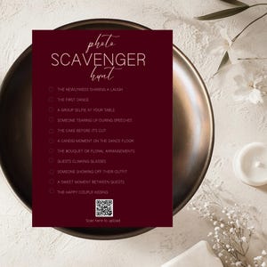 Puede incluir: Una tarjeta borgoña de búsqueda del tesoro fotográfica con una lista de indicaciones, incluyendo "Los recién casados compartiendo una risa" y "El primer baile". La tarjeta está sobre un plato de color bronce, con un código QR en la parte inferior. El texto "búsqueda del tesoro fotográfica" está escrito en una fuente decorativa.
