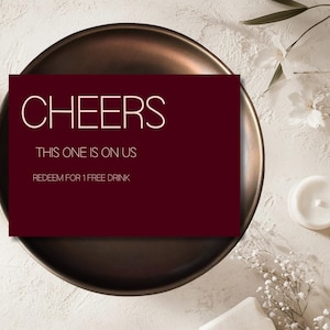 May include: A burgundy card with the word "CHEERS" in white, along with the phrases "THIS ONE IS ON US" and "REDEEM FOR 1 FREE DRINK". The card rests on a bronze-colored plate, with floral and candle accents.