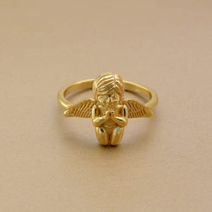 May include: A gold-colored ring featuring a small angel with folded hands and outstretched wings. The angel is kneeling, and the ring band is a simple, smooth design. The ring is set against a neutral background.