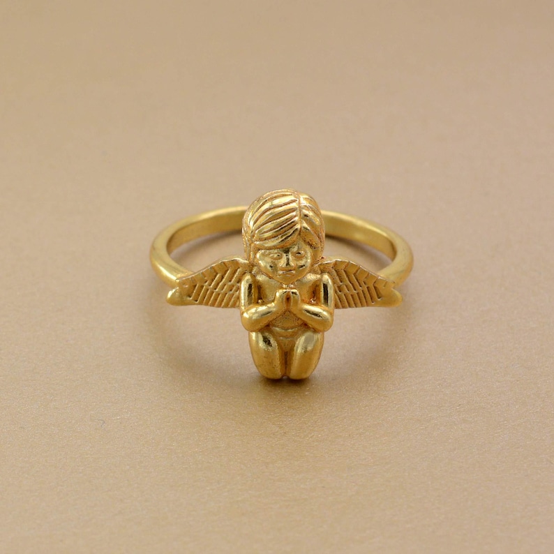 May include: A gold-colored ring featuring a small angel with folded hands and outstretched wings. The angel is kneeling, and the ring band is a simple, smooth design. The ring is set against a neutral background.