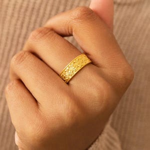 May include: A gold-colored ring with a detailed floral pattern. The ring is worn on a finger, showcasing its intricate design and bright, shiny finish. The ring's band is approximately 0.5 inches wide.