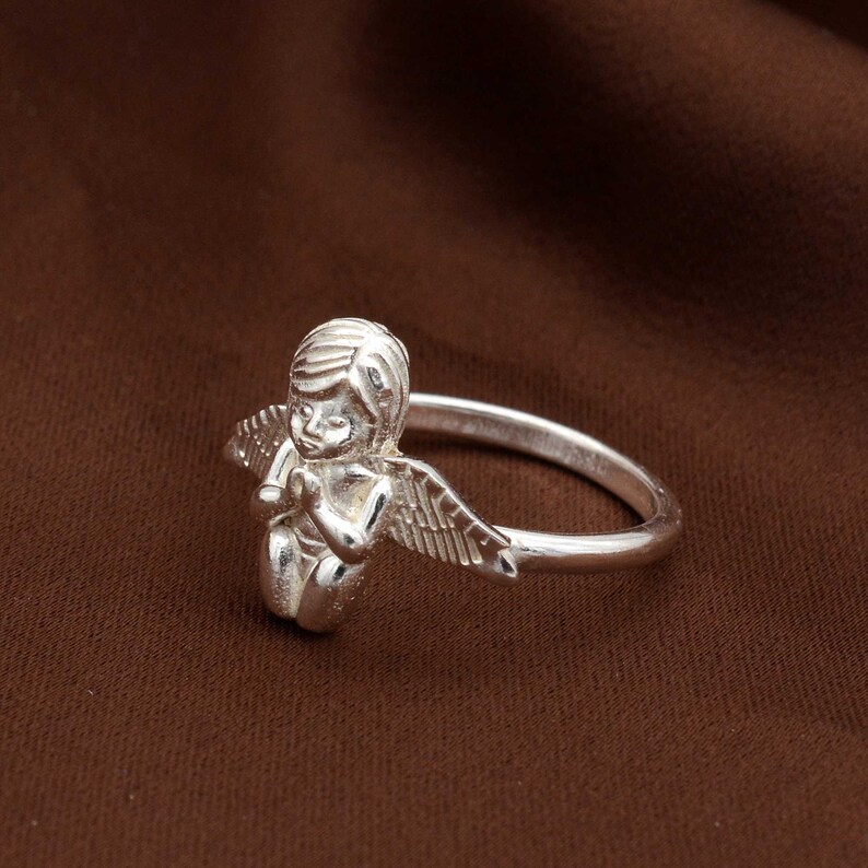 May include: A silver ring featuring a detailed angel figurine with outstretched wings. The angel is kneeling with hands clasped in prayer. The ring band is a simple, polished silver. The jewelry is set against a brown background.