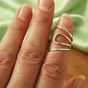 May include: A silver-colored ring with a unique, flowing design. The ring wraps around the finger with multiple curved bands, creating an artistic and modern look. The jewelry is set against a soft green background.