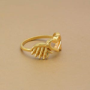 May include: A gold-colored ring featuring a heart-shaped design and a decorative wing element. The ring is made of a shiny, metallic material and is set against a neutral background. The ring's design is detailed and intricate.