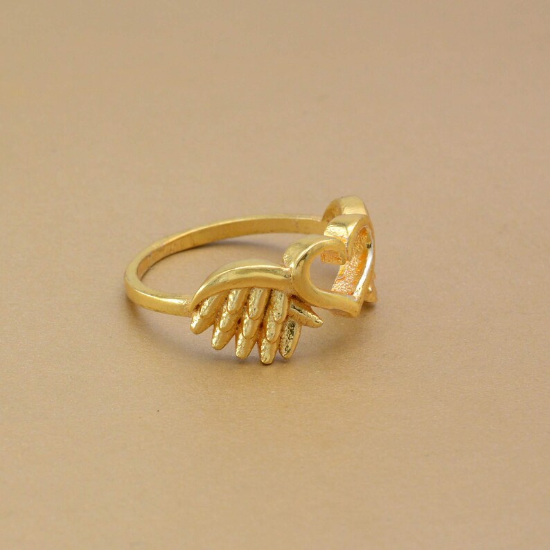 May include: A gold-colored ring featuring a heart-shaped design and a decorative wing element. The ring is made of a shiny, metallic material and is set against a neutral background. The ring's design is detailed and intricate.