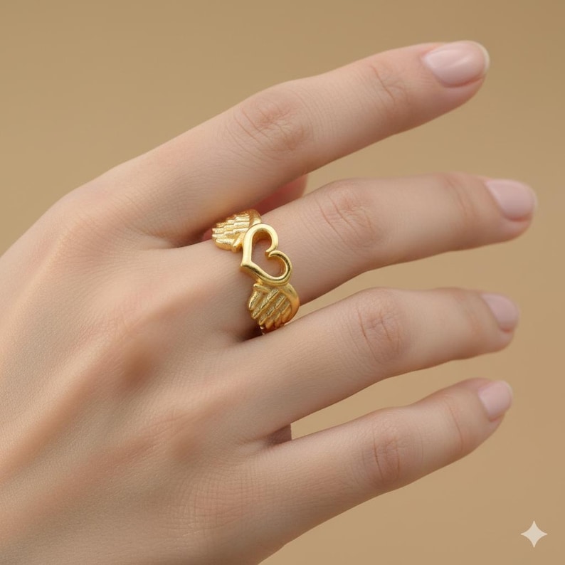 May include: A gold-colored ring featuring a heart-shaped design with wing accents. The ring is worn on a finger, showcasing its intricate details and metallic sheen. The ring's design is a heart shape with wings on each side.