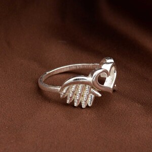 May include: A silver ring featuring a heart and wing design. The ring has a polished finish and is set against a brown background. The heart is open, and the wings are detailed.