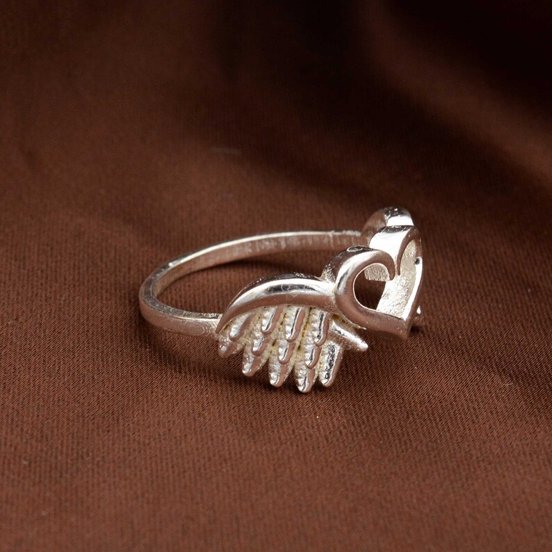 May include: A silver ring featuring a heart and wing design. The ring has a polished finish and is set against a brown background. The heart is open, and the wings are detailed.