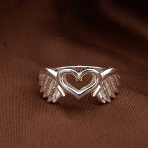 May include: A silver ring featuring a heart-shaped design with angel wings on either side. The ring is made of a shiny metal and is set against a brown background. The ring's design is detailed and intricate, with a focus on the heart and wing shapes.