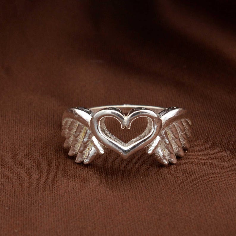 May include: A silver ring featuring a heart-shaped design with angel wings on either side. The ring is made of a shiny metal and is set against a brown background. The ring's design is detailed and intricate, with a focus on the heart and wing shapes.